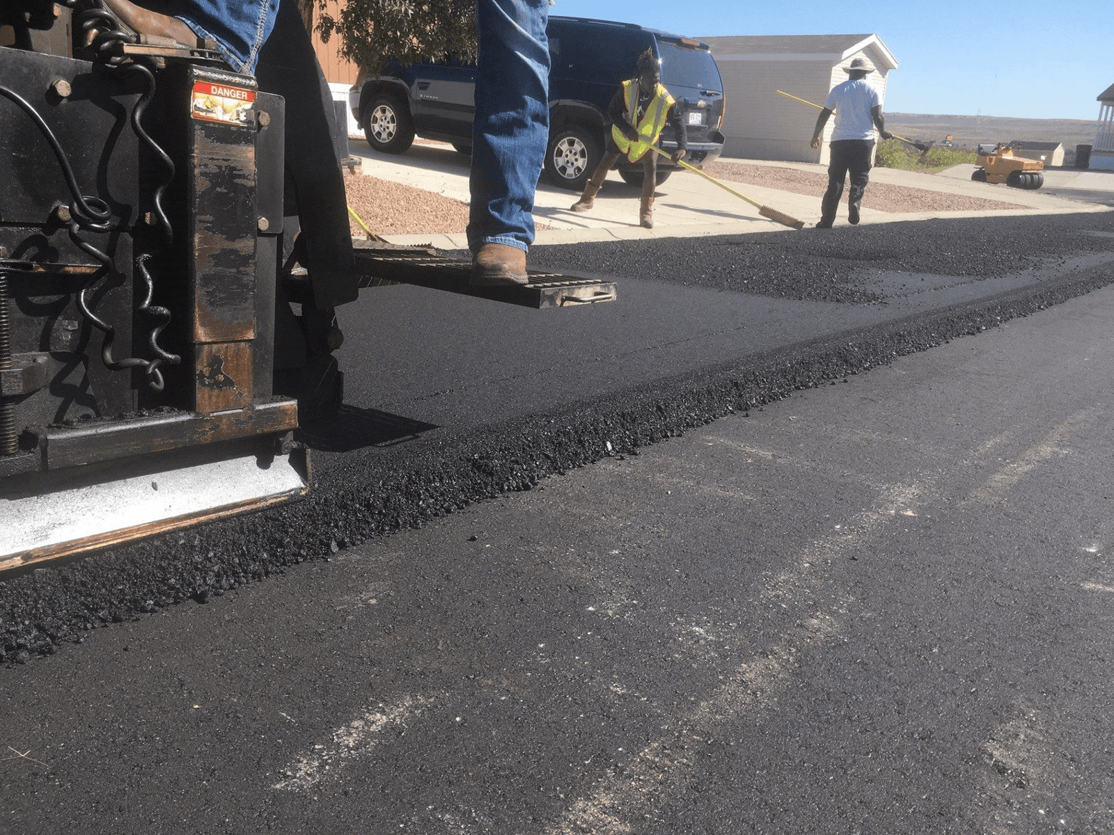 An HEV worker beside the paving machine laying a fresh ribbon of hot-mix asphalt on a residential street