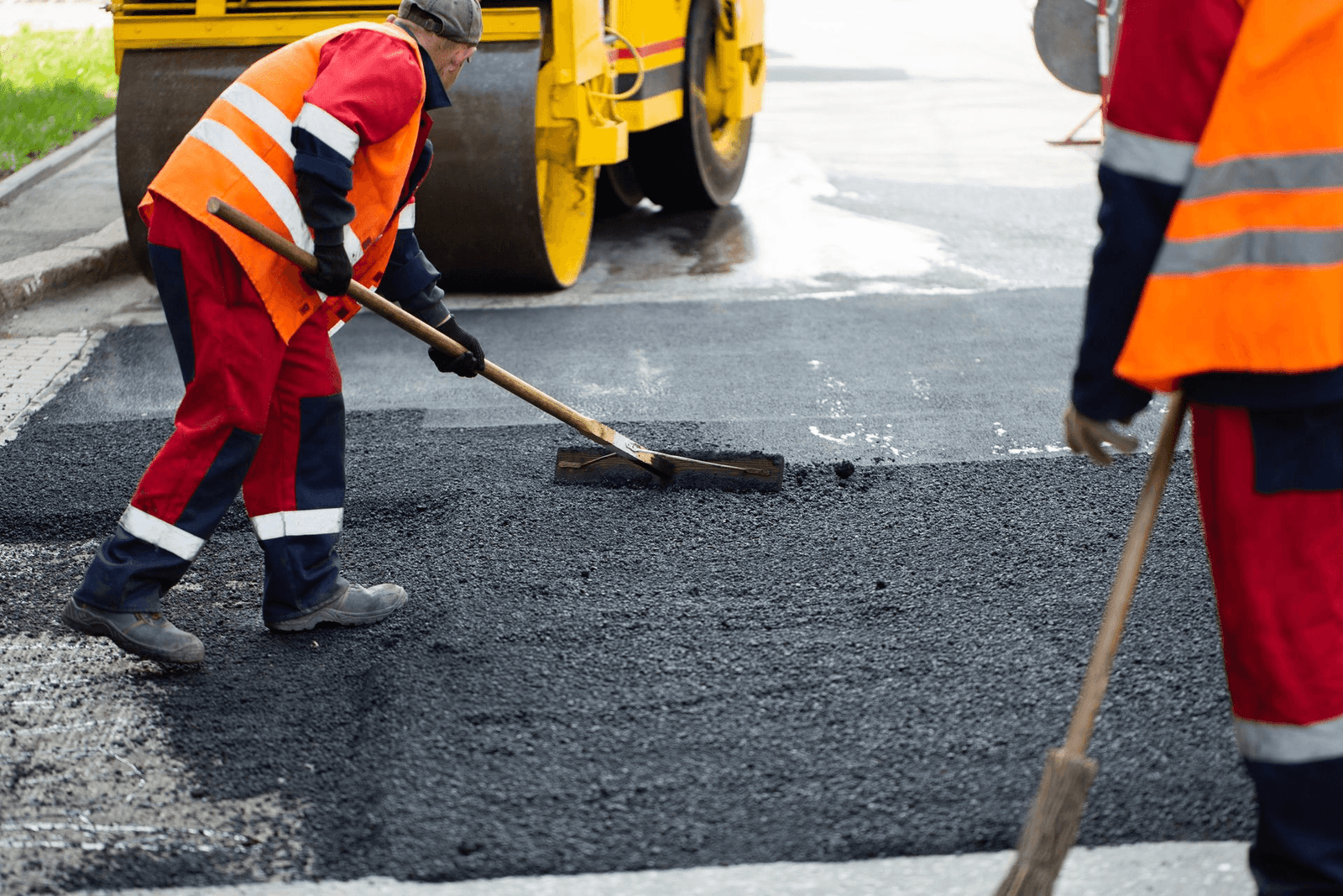HEV crew in hi-vis spreading and raking fresh hot-mix asphalt behind a roller on site